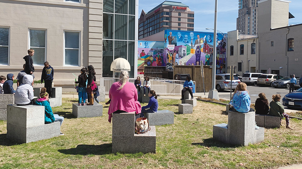 Students and teacher sitting outside, with a large colorful mural visible in far distance