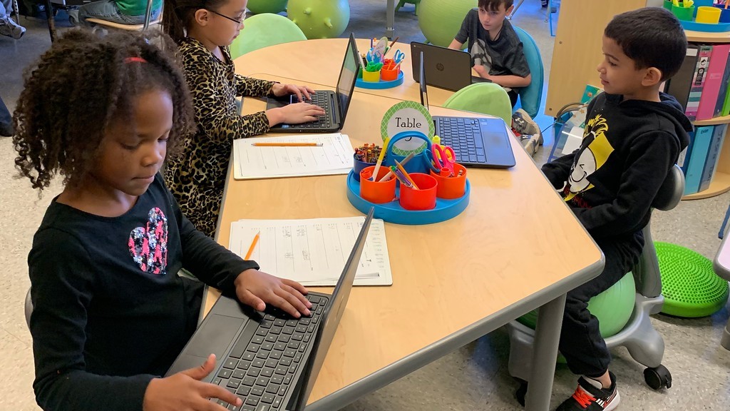 a young student works on a laptop at a table with her classmates