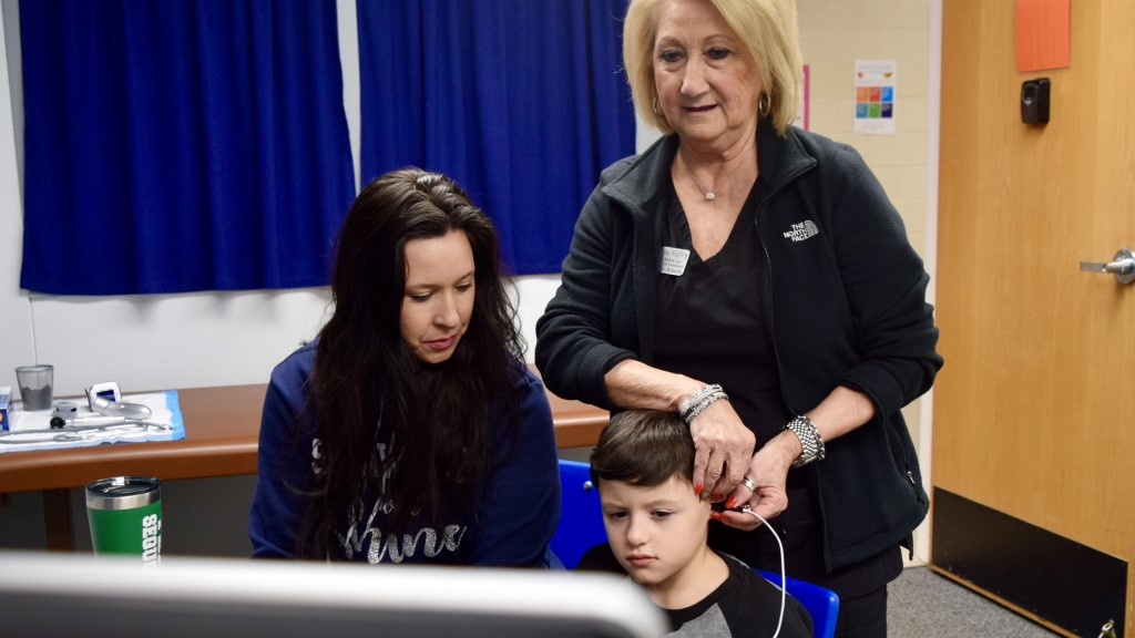 An adult performs an ear examine on a young student with his guardian present