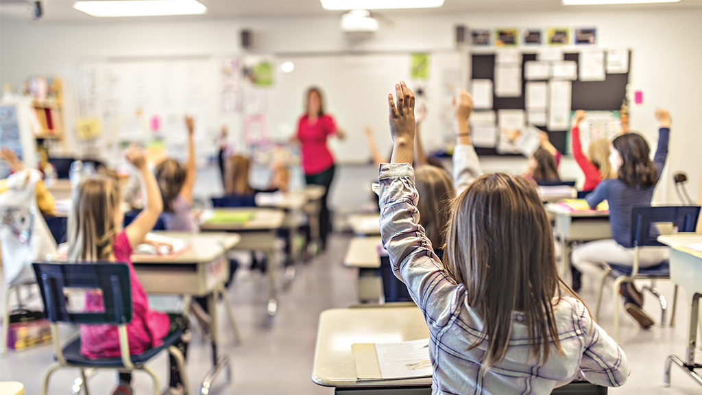 a picture taken at the back of a classroom. the student closest to the camera raises her hand