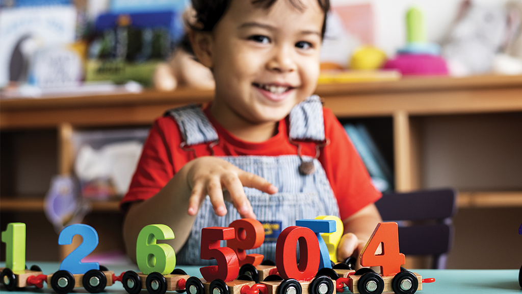 a boy, in the background, smiles at the camera. he is playing with wood numbers that are in the photograph's foreground 