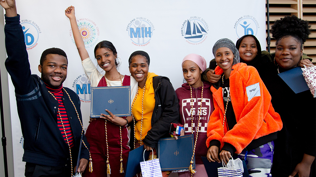 a group of students celebrate receiving degrees, with honor cords around their neck. the students are joyful, two holding up their hands, and a few embracing