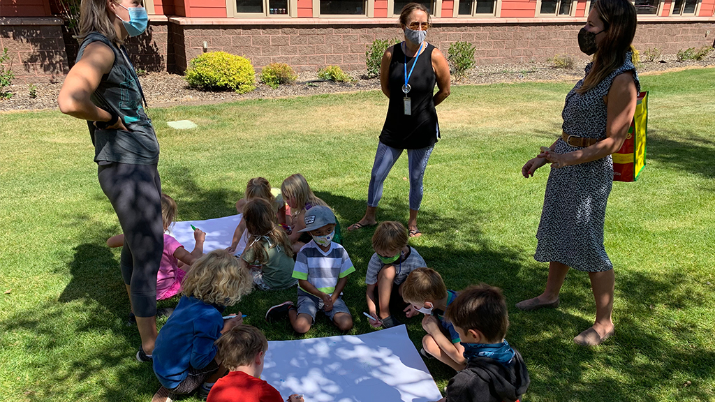 a group of students with masks sit on the grass and do an activity while their three teachers in masks talk