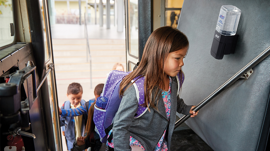 a student climbs up the steps of a school bus, with a purell hand sanitizer mounted to the interior of the bus 