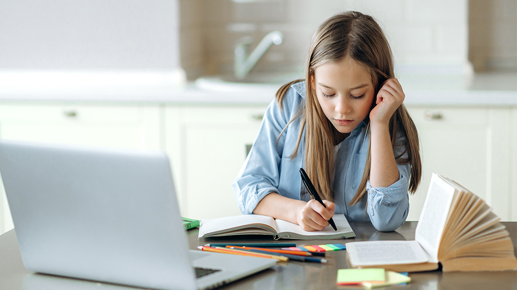 a girl writes in a notebook with a laptop nearby