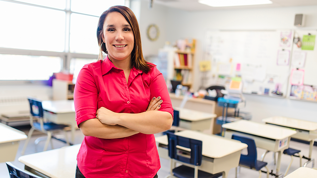 A teacher stands with her arms folded across her chest in an empty classroom. 