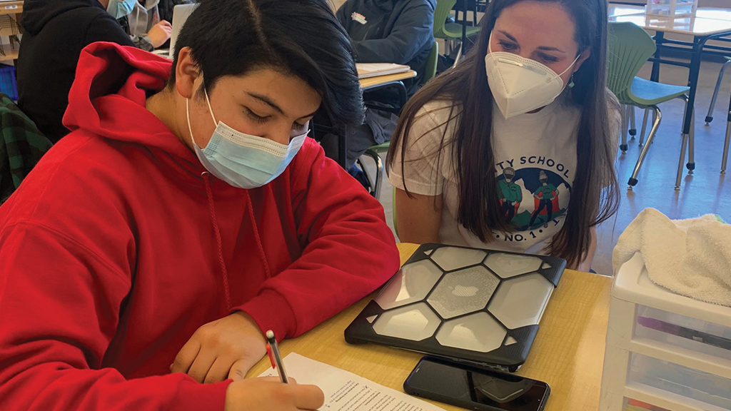 A teacher watches a high school student write on a piece of paper.