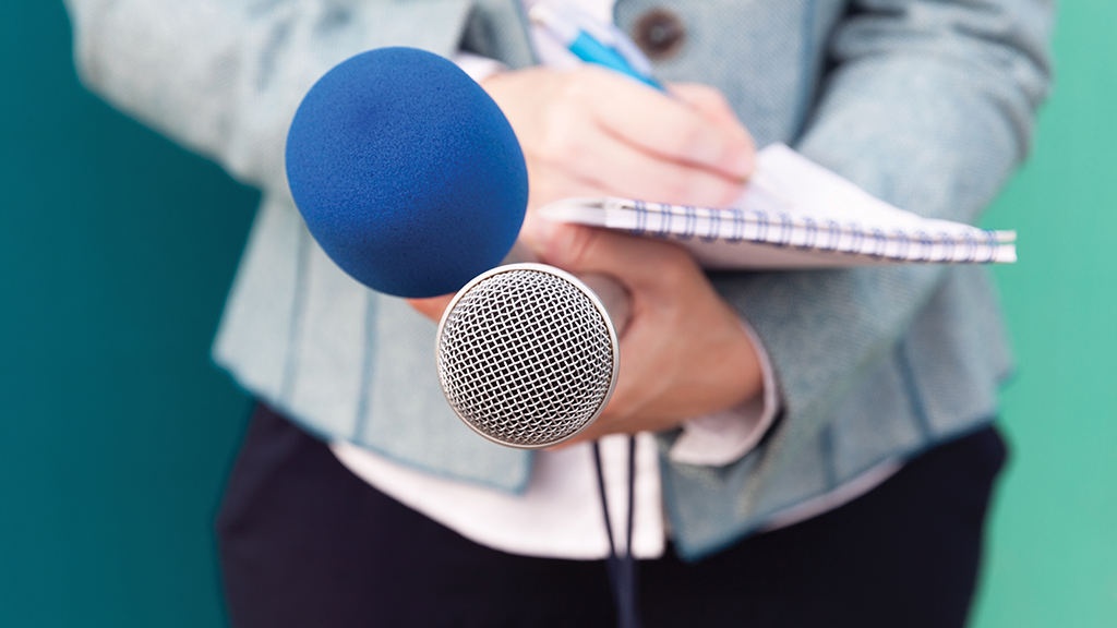 A female news reporter stands with a microphone, notebook and tape recorder in her hand