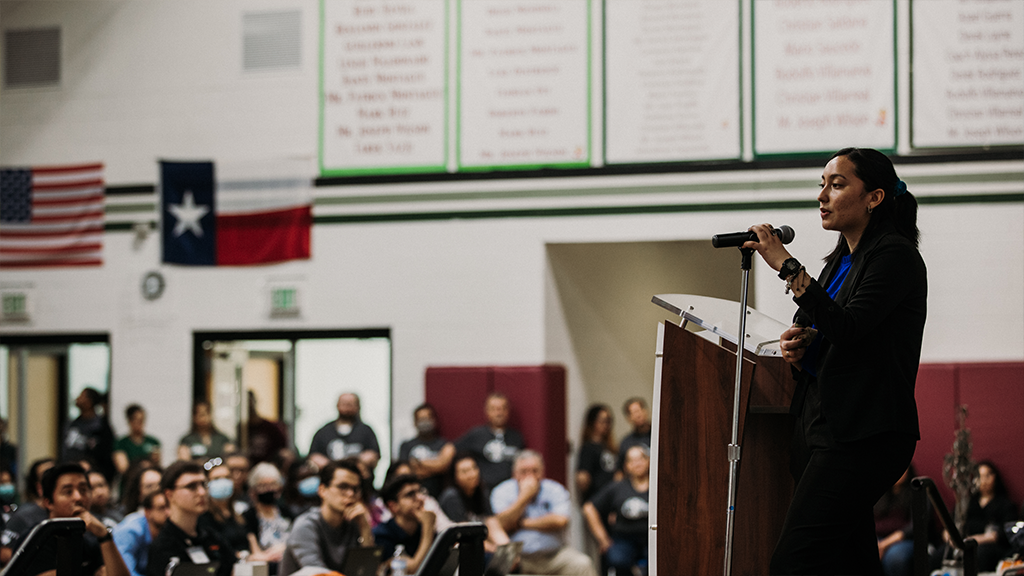 A high school student stands before a podium and speaks to a large audience about her science project.