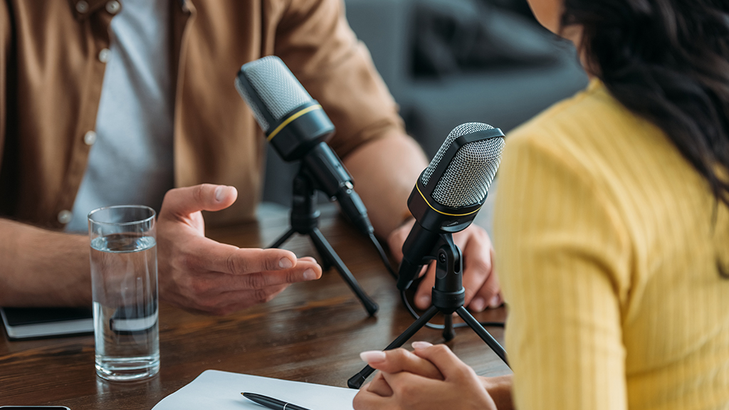 A man and a woman sit at microphones on a desk and participate in a podcast.