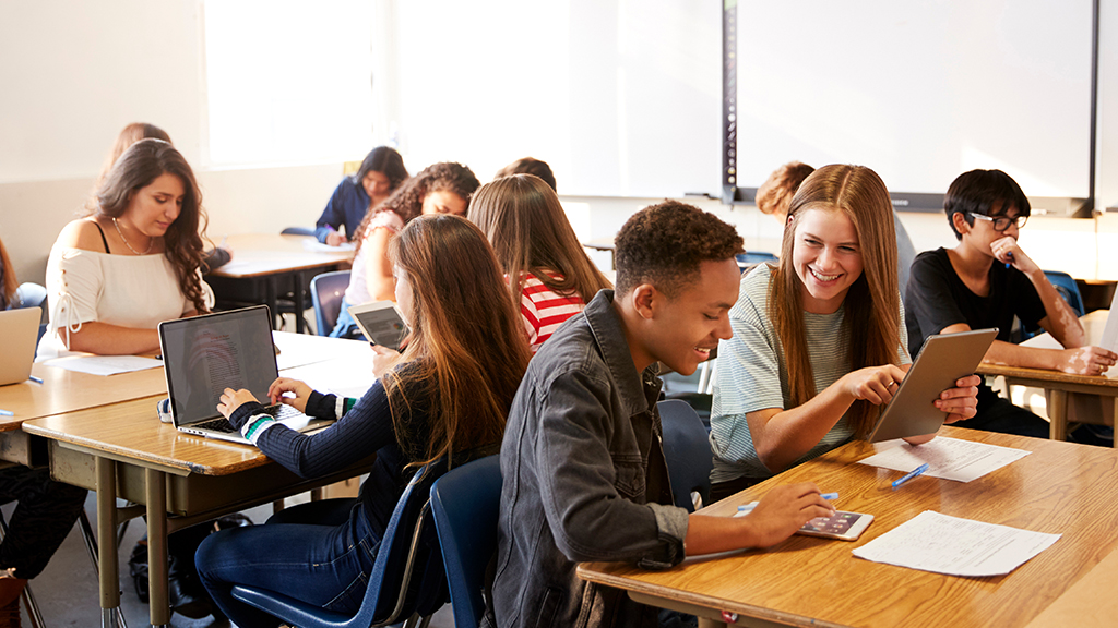 A group of high school-age students talk and work in small groups while seated at their classroom desks. 