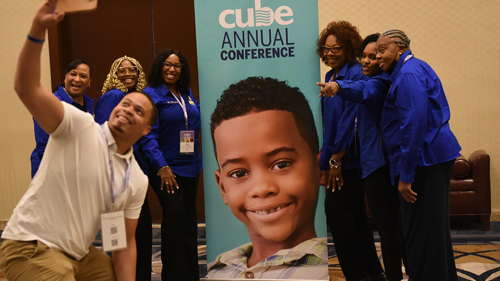 A group of smiling conference goers talk a self-portrait in front of a conference poster.