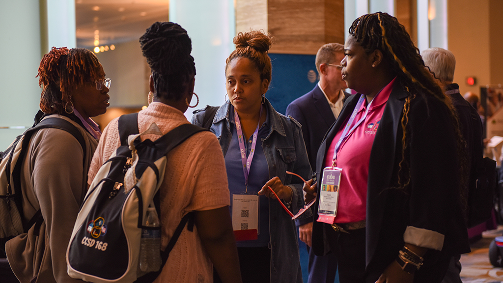 Four women wear backpacks and with papers in their hands, stand and talk during a break during a conference meeting.