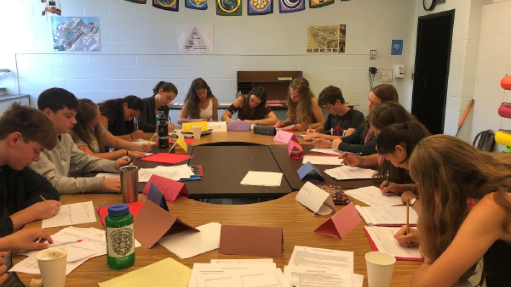 A group of high school students sit around a round table as they participate in a class. 