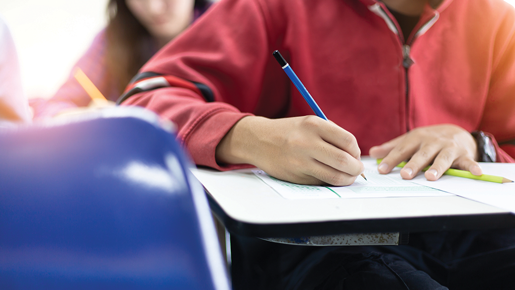 the hands of a teenage boy are seen as he writes in a booklet