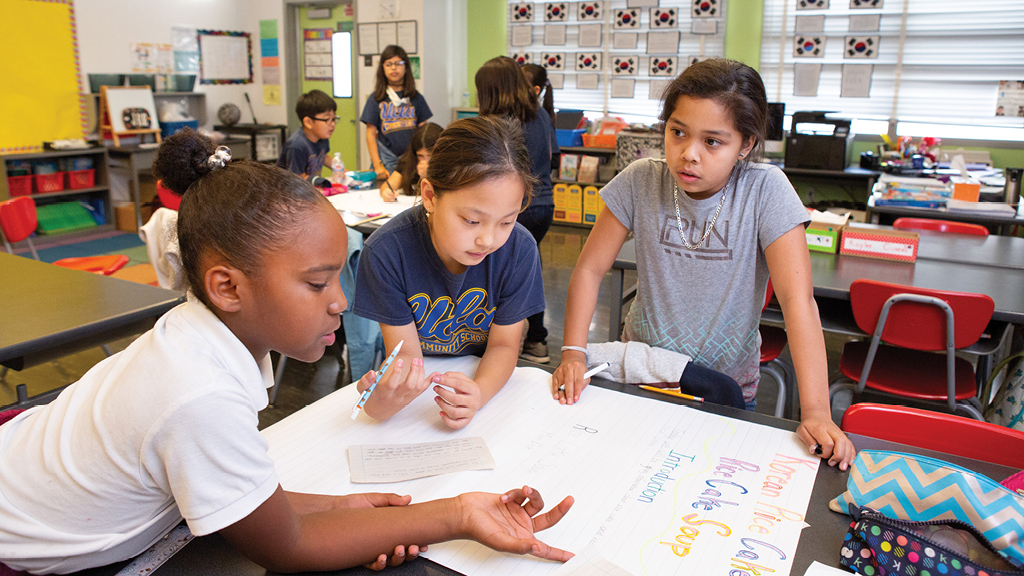 Three elementary-age students do work together at a school desk. 