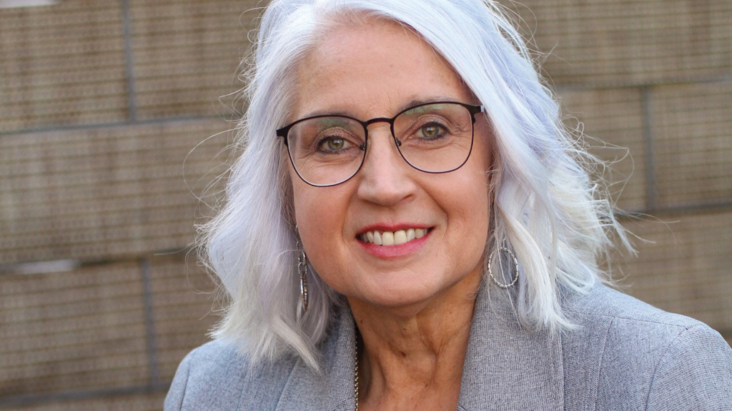 A woman with silver hair and wearing eyeglasses smiles as she looks into the camera.