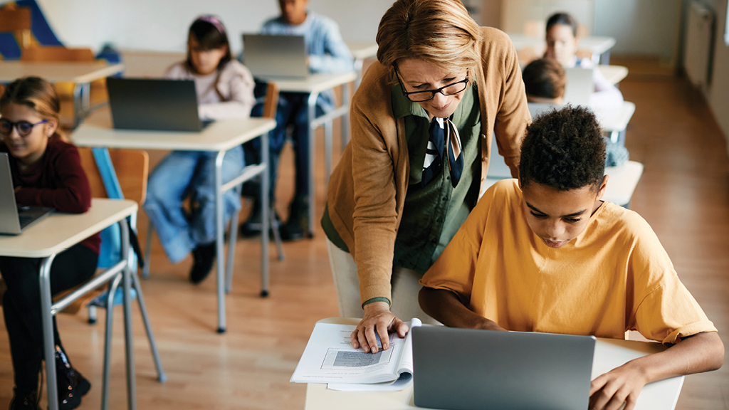 A teacher looks over the shoulder of a student as he works on a laptop.