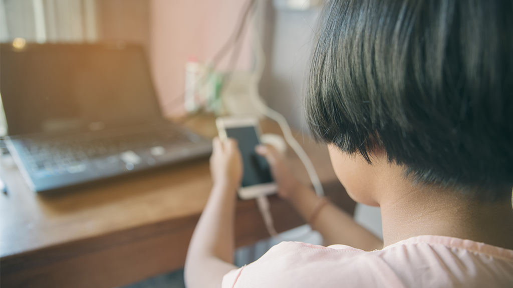 a student, only shown from the back, is using a mobile phone while a laptop sits on a desk in front of her